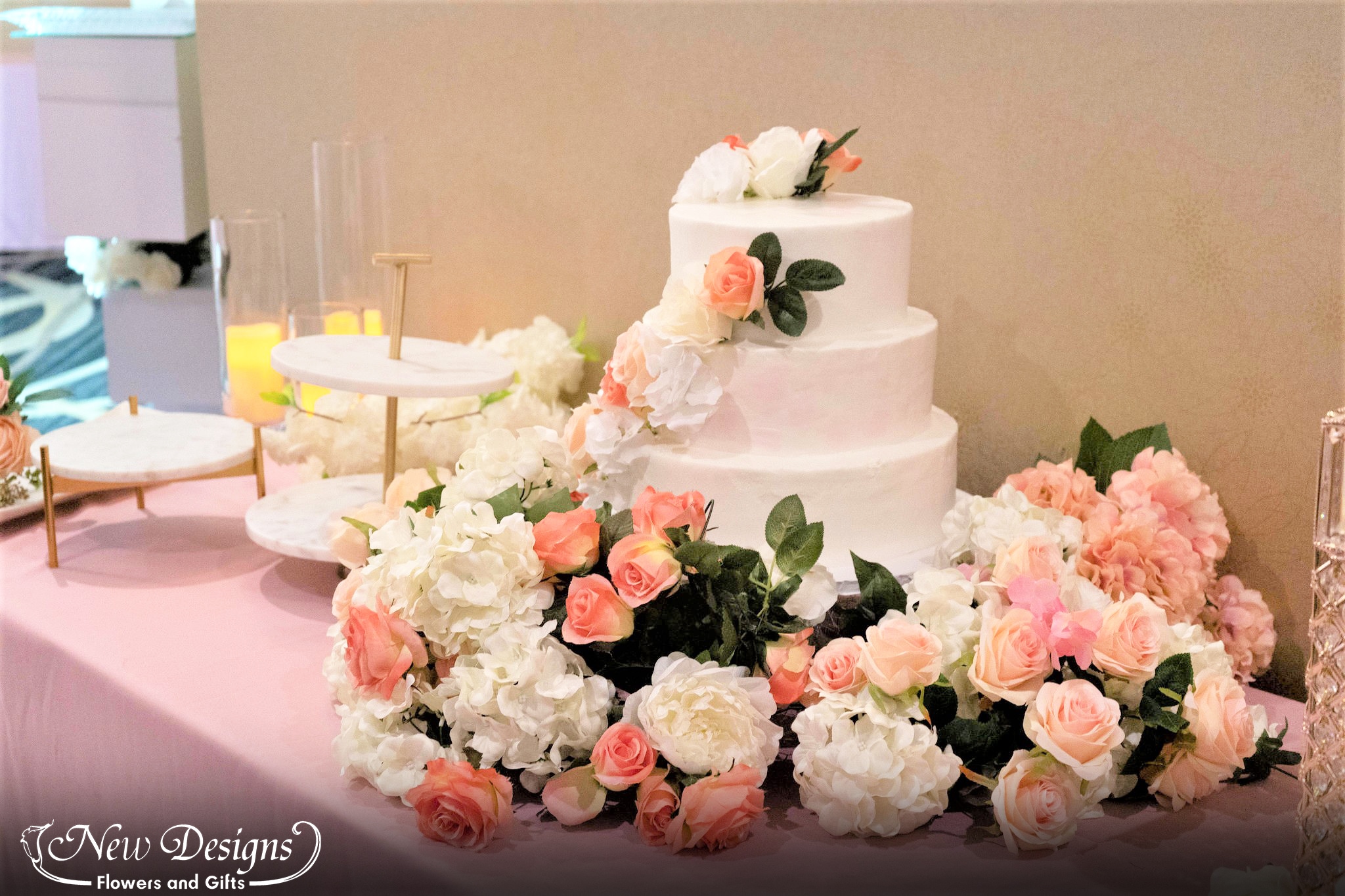 A white cake wrapped and overflowing with white neutral and orange roses, sits on top of a dusty pink tablecloth,